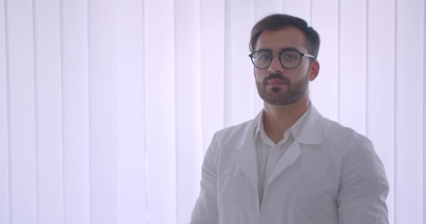 Closeup retrato de adulto bonito branco médico masculino em óculos olhando para a câmera sorrindo alegremente na sala branca dentro de casa — Vídeo de Stock