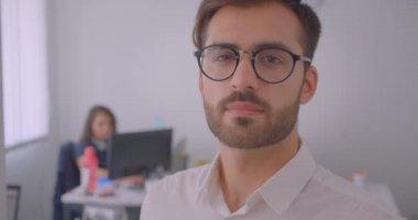 Yakışıklı başarılı kafkas işadamının gözlüklü yakın çekim portresi arka planda çalışanla birlikte kapalı alanda beyaz ofisteki kameraya bakıyor