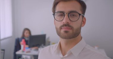 Yakışıklı başarılı beyaz işadamının gözlüklü yakın çekim portresi arka planda çalışanla birlikte kapalı alanda beyaz ofisteki kameraya bakıyor