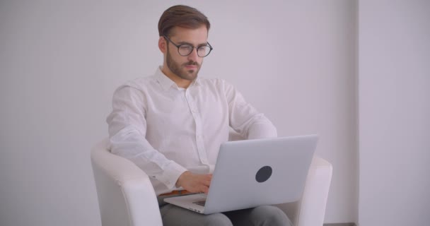 Close-up portret van volwassen knappe bebaarde Kaukasische zakenman in glazen met behulp van de laptop zittend in de fauteuil in het witte kantoor binnenshuis — Stockvideo