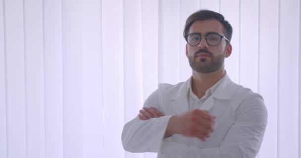 Gözlüklü yetişkin yakışıklı sakallı beyaz erkek doktorun yakın çekim portresi kollarını göğsüne doladı kapalı beyaz odadaki kameraya bakarak — Stok video