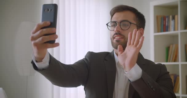 Closeup portrait of adult handsome bearded caucasian businessman in glasses having a video call on the phone in the office indoors with bookshelves on the background — Stock Video