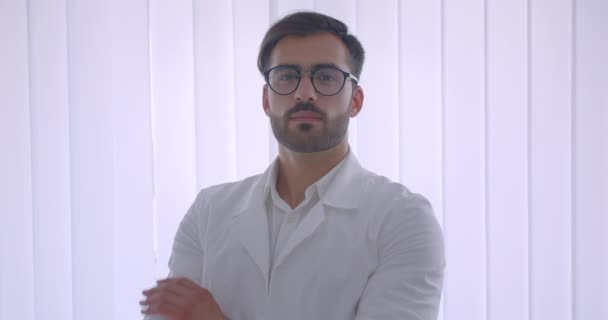 Zbliżenie portret dorosłego przystojnego kaukaski mężczyzna lekarz w okularach z rękami przekreślone klatki piersiowej patrząc na kamerę w białym pomieszczeniu w pomieszczeniu — Wideo stockowe