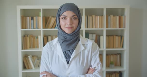 Primo piano ritratto di giovane musulmana attraente medico donna in hijab guardando la fotocamera sorridente felicemente con le braccia incrociate sul petto in biblioteca al chiuso — Video Stock