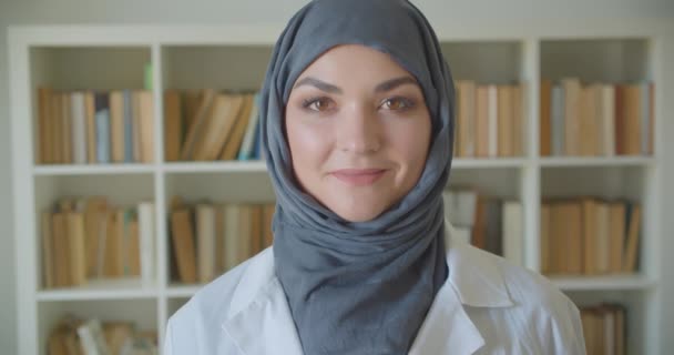 Nahaufnahme Porträt einer jungen muslimischen attraktiven Ärztin im Hijab, die glücklich lächelnd in die Kamera blickt, während sie in der Bibliothek drinnen steht — Stockvideo