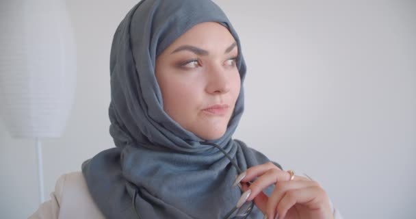 Retrato de close-up de jovem muçulmano atraente médico feminino em hijab e casaco branco olhando para câmera segurando óculos em consideração sentado na poltrona na sala branca dentro de casa — Vídeo de Stock