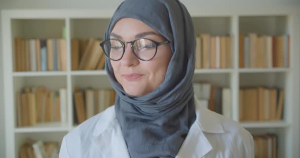 Zbliżenie portret młodego muzułmańskiego atrakcyjny żeński lekarz w hidżab i okulary patrząc na aparat uśmiechnięty wesoło stojący w bibliotece w pomieszczeniu — Wideo stockowe