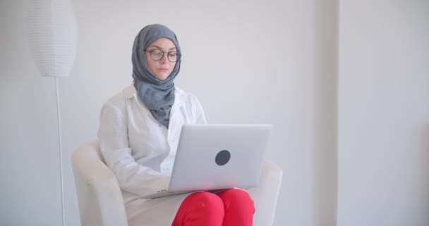 Retrato de cerca de la joven musulmana atractiva doctora en hijab y bata blanca usando el portátil mirando a la cámara sonriendo felizmente sentada en el sillón en la habitación blanca en el interior — Vídeo de stock