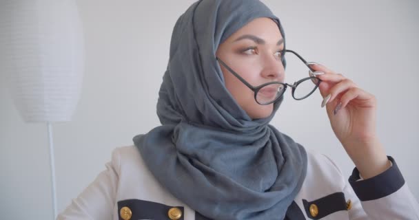 히잡과 흰색 코트를 입은 젊은 무슬림 매력적인 여성 의사의 클로즈업 초상화가 실내의 흰색 방에 안락의자에 앉아 안경을 들고 카메라를 바라보고 있습니다. — 비디오