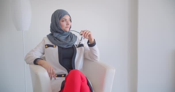 Closeup portrait of young muslim attractive female doctor in hijab and white coat looking at camera holding eyeglasses thoughtfully sitting in the armchair in the white room indoors — Stock Video