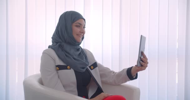 Κοντινά πλάνα πορτραίτο του νεαρού μουσουλμανικού γυναικείου γιατρού σε χιτζάμπ και λευκό παλτό με βιντεοκλήση στο tablet που κάθεται στην πολυθρόνα στο λευκό δωμάτιο σε εσωτερικούς χώρους — Αρχείο Βίντεο