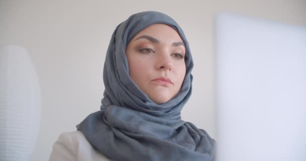Nahaufnahme Porträt einer jungen attraktiven muslimischen Ärztin in Hijab und weißem Mantel, die auf dem Laptop tippt und glücklich lächelnd in die Kamera blickt, im Sessel im weißen Raum drinnen — Stockvideo