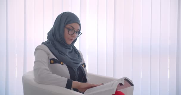 特写侧看年轻穆斯林有吸引力的女医生在头巾和白色外套读一本书看着相机微笑愉快地坐在室内白色房间的扶手椅 — 图库视频影像