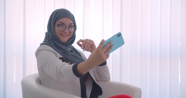 히잡과 흰색 코트를 입은 젊은 무슬림 여성 의사의 클로즈업 사이드 뷰 초상화가 실내의 흰색 방에 안락의자에 유쾌하게 앉아 미소짓고있는 휴대 전화에서 셀카를 찍고 있습니다. — 비디오