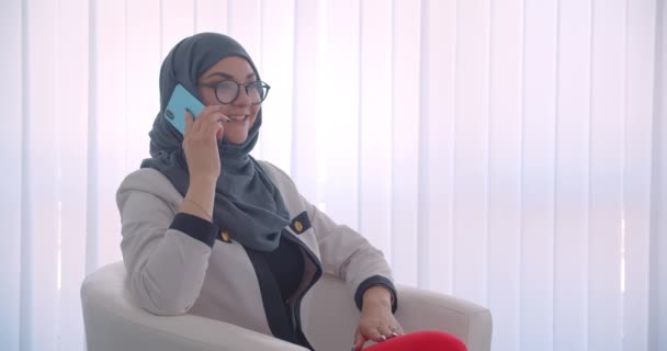 Closeup oldalnézetből portréja fiatal muzulmán vonzó női orvos a hidzsáb és fehér kabát, amelynek telefonhívást ül a karosszékben a fehér szobában beltéri — Stock videók