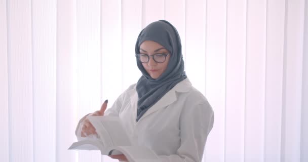 Visão lateral close-up retrato de jovem muçulmano atraente médico feminino em hijab e casaco branco lendo um livro olhando para a câmera sorrindo alegremente em pé na sala branca dentro de casa — Vídeo de Stock