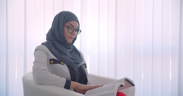 Primo piano vista laterale ritratto di giovane musulmana attraente medico donna in hijab e cappotto bianco leggere un libro seduto nella poltrona nella stanza bianca all'interno — Video Stock