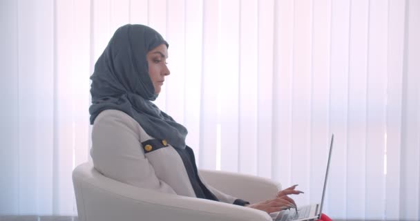 Nahaufnahme Seitenansicht Porträt einer jungen muslimischen attraktiven Ärztin im Hijab und weißem Mantel, die auf dem Laptop im Sessel im weißen Raum im Innenraum sitzt — Stockvideo