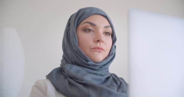 Retrato de cerca de la joven musulmana atractiva doctora en hijab y bata blanca escribiendo en el portátil mirando a la cámara sonriendo alegremente sentada en el sillón en la habitación blanca en el interior — Vídeos de Stock