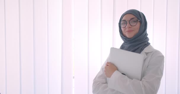 Primer plano vista lateral retrato de joven musulmana atractiva doctora en hijab y abrigo blanco sosteniendo un ordenador portátil mirando a la cámara de pie en la habitación blanca en el interior — Vídeos de Stock