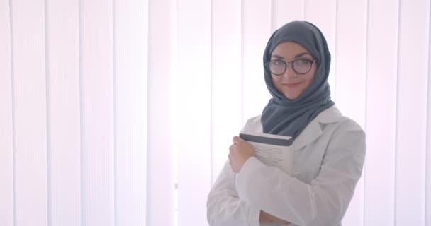 特写侧看年轻穆斯林有吸引力的女医生在头巾和白色外套拿着一本书看着相机微笑愉快地站在室内的白色房间的肖像 — 图库视频影像