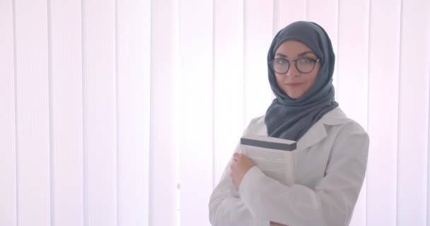 Nahaufnahme Seitenansicht Porträt einer jungen Muslimin attraktive Ärztin in Hijab und weißem Mantel hält ein Buch im weißen Raum im Haus stehen — Stockvideo