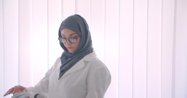 Visão lateral close-up retrato de jovem muçulmano atraente médico feminino em hijab e casaco branco lendo um livro em pé na sala branca dentro de casa — Vídeo de Stock