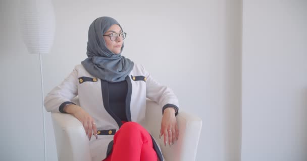 Gros plan portrait de jeune musulmane séduisante médecin en hijab et manteau blanc regardant la caméra souriant joyeusement assis dans le fauteuil dans la chambre blanche à l'intérieur — Video