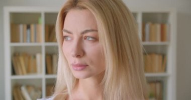 Arka planda kitaplık ile kütüphanede mutlu bir şekilde gülümseyen kamera görünümlü yetişkin beyaz sarışın kadın çekim portre