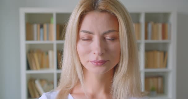 Gros plan portrait d'adulte caucasien blonde femelle regardant la caméra souriant joyeusement à l'intérieur dans la bibliothèque avec des étagères sur le fond — Video