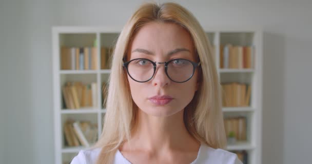Close-up portret van volwassen Kaukasische blonde vrouw in glazen kijken naar camera binnenshuis in de bibliotheek — Stockvideo
