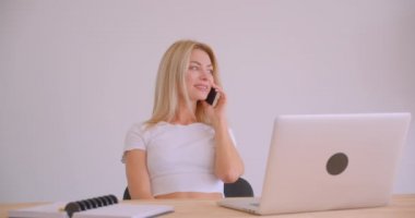 Masa başında ofiste telefonda kapalı neşeli bir konuşma sahip laptop kullanarak yetişkin beyaz sarışın işkadını portre kapatmak