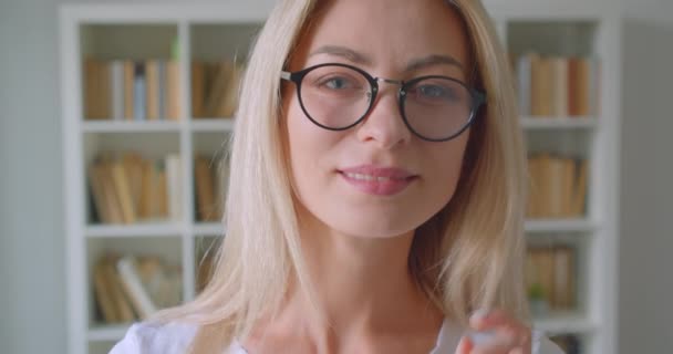 Gros plan portrait d'adulte caucasien blonde femelle dans des lunettes regardant la caméra souriant joyeusement à l'intérieur dans la bibliothèque — Video