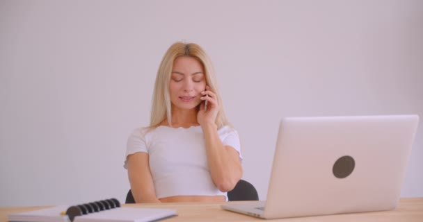 Nahaufnahme Porträt einer erwachsenen kaukasischen blonden Geschäftsfrau mit dem Laptop, die drinnen im Büro am Schreibtisch telefoniert — Stockvideo