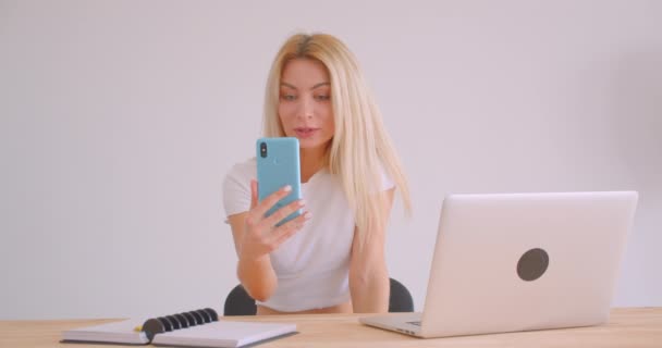 Gros plan portrait d'adulte caucasien blonde femme d'affaires ayant un appel vidéo sur le téléphone assis devant l'ordinateur portable à l'intérieur dans le bureau — Video