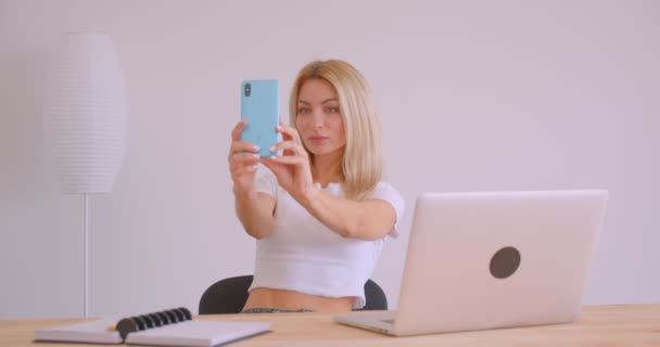 オフィスの屋内でラップトップの前に座って携帯電話にセルフを取る大人の白人ブロンドのビジネスウーマンのクローズアップ肖像画 — ストック動画