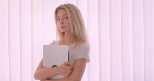 Closeup portrait of adult caucasian blonde businesswoman holding the laptop looking at camera smiling happily standing indoors in the office — Stock Video