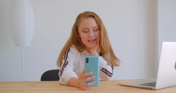 Крупный план портрета молодой красивой кавказской девушки с видеозвонком по телефону перед ноутбуком в помещении в квартире — стоковое видео