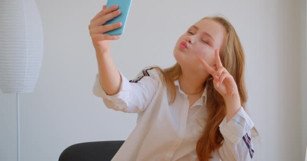 아파트에서 노트북 앞에 앉아 포즈를 취하는 전화에 셀카를 찍는 젊은 예쁜 백인 소녀의 클로즈업 초상화 — 비디오