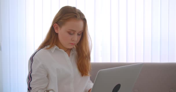 Retrato de close-up de jovem menina bonita caucasiana usando o laptop olhando para a câmera sorrindo feliz sentado no sofá dentro de casa no apartamento — Vídeo de Stock