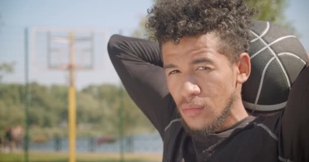 Closeup retrato de jovem afro-americano bonito jogador de basquete masculino olhando para a câmera segurando uma bola atrás de suas costas ao ar livre — Vídeo de Stock