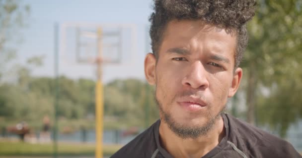 Närbild porträtt av unga stilig afrikansk amerikansk manlig basketspelare tittar på kamera med allvarliga uttryck utomhus — Stockvideo