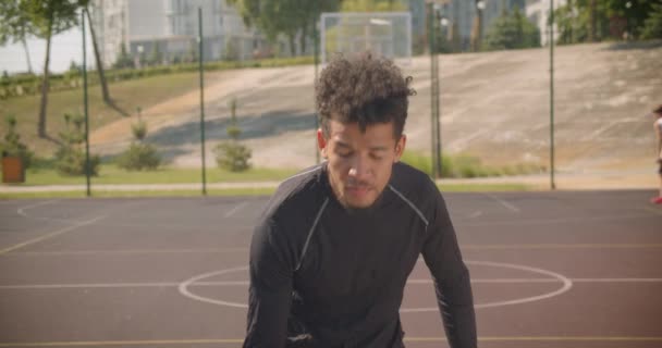 Retrato de primer plano del joven guapo jugador de baloncesto afroamericano lanzando una pelota en un aro al aire libre con gente en el fondo — Vídeos de Stock