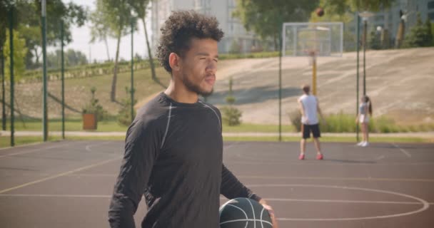 Крупним планом портрет молодого красивого афроамериканського баскетболіста, який тримає м'яч і дивиться на камеру на відкритому повітрі — стокове відео