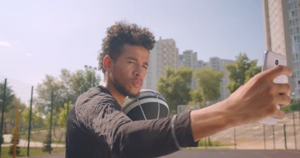 Крупный план портрета молодого спортивного африканского баскетболиста мужского пола, держащего мяч и делающего селфи по телефону на улице на площадке со зданиями на заднем плане — стоковое видео
