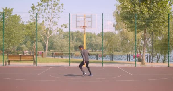 Retrato de primer plano del joven jugador de baloncesto afroamericano activo lanzando una pelota en un aro al aire libre en la cancha — Vídeos de Stock
