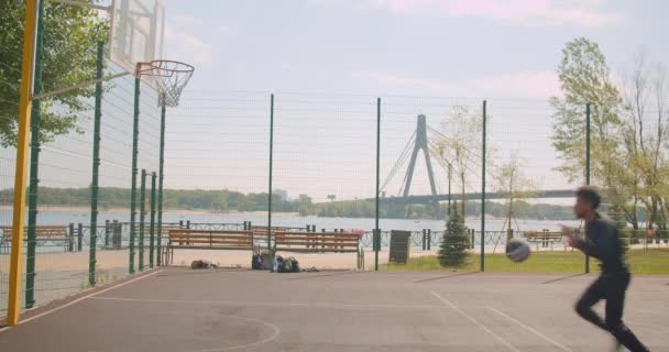 スポーティーな魅力的なアフリカ系アメリカ人男性バスケットボール選手の肖像画は、背景に橋と都市の屋外のコートにフープにボールを投げる — ストック動画