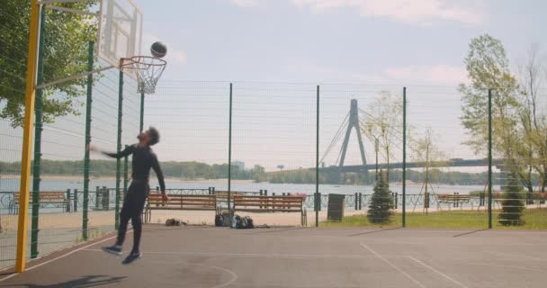 Retrato de deportista atractivo afroamericano jugador de baloncesto masculino lanzando una pelota en un aro y celebrando con triunfo en la cancha en la ciudad urbana al aire libre con puente en el fondo — Vídeo de stock