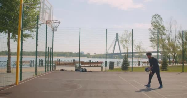 Retrato de deportista feliz afroamericano jugador de baloncesto masculino lanzando una pelota en un aro y celebrando con triunfo en la cancha en la ciudad urbana al aire libre con puente en el fondo — Vídeo de stock