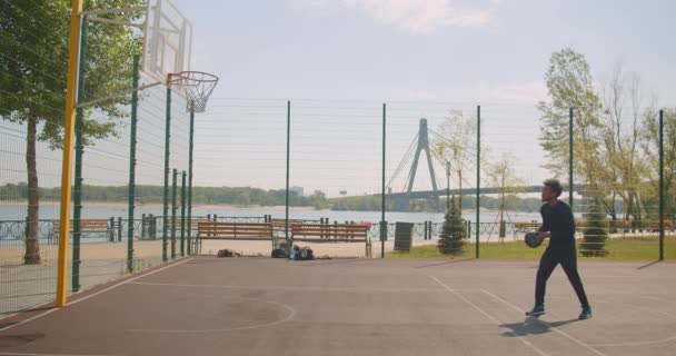 Retrato de deportista atractivo afroamericano jugador de baloncesto masculino lanzando una pelota en un aro y no ser infeliz en la cancha en la ciudad urbana al aire libre con puente en el fondo — Vídeo de stock